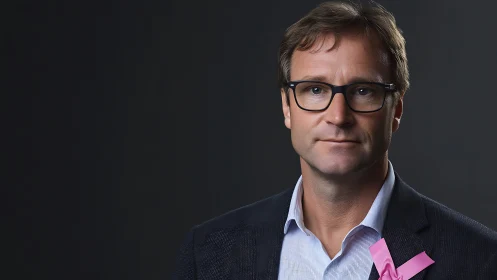 Middle aged man in suit with pink ribbon on dark backdrop.