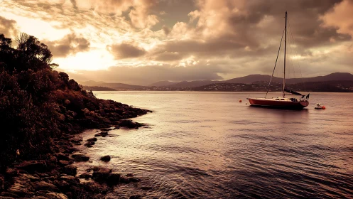 Sunset sailboat anchored off rocky coastal shoreline.
