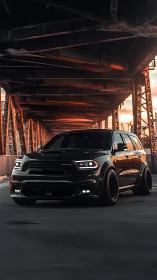 Widebody black SUV under steel bridge at sunset glow.