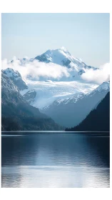 Snowcapped mountain above calm lake with low white clouds.