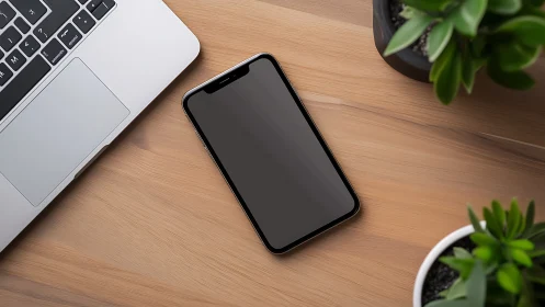 Smartphone lies face up on wooden desk beside laptop and plants
