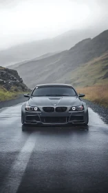 Matte black BMW coupe on wet mountain road in misty rain.
