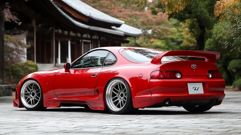 Red Toyota Supra stands lowered beside tranquil Japanese temple.