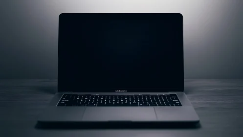 Open laptop with dark screen on plain surface in low light.