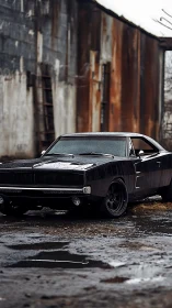 Black classic muscle car in wet industrial yard setting.