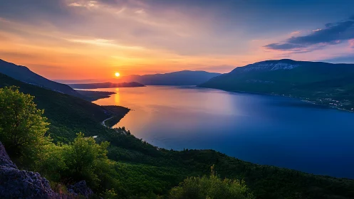 Sunset over calm blue lake with surrounding green hills.