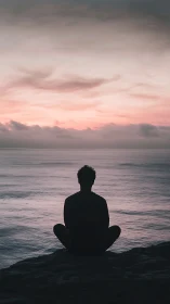 Silhouette meditates by ocean under pastel dawn sky.