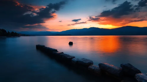 Rock jetty on calm lake under deep orange sunset sky.