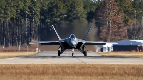 Stealth fighter jet powers down runway against forest backdrop.