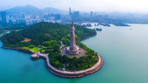 Towering coastal statue amid green peninsula and skyline.