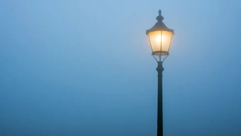 Solitary streetlamp glows softly through dense morning fog.